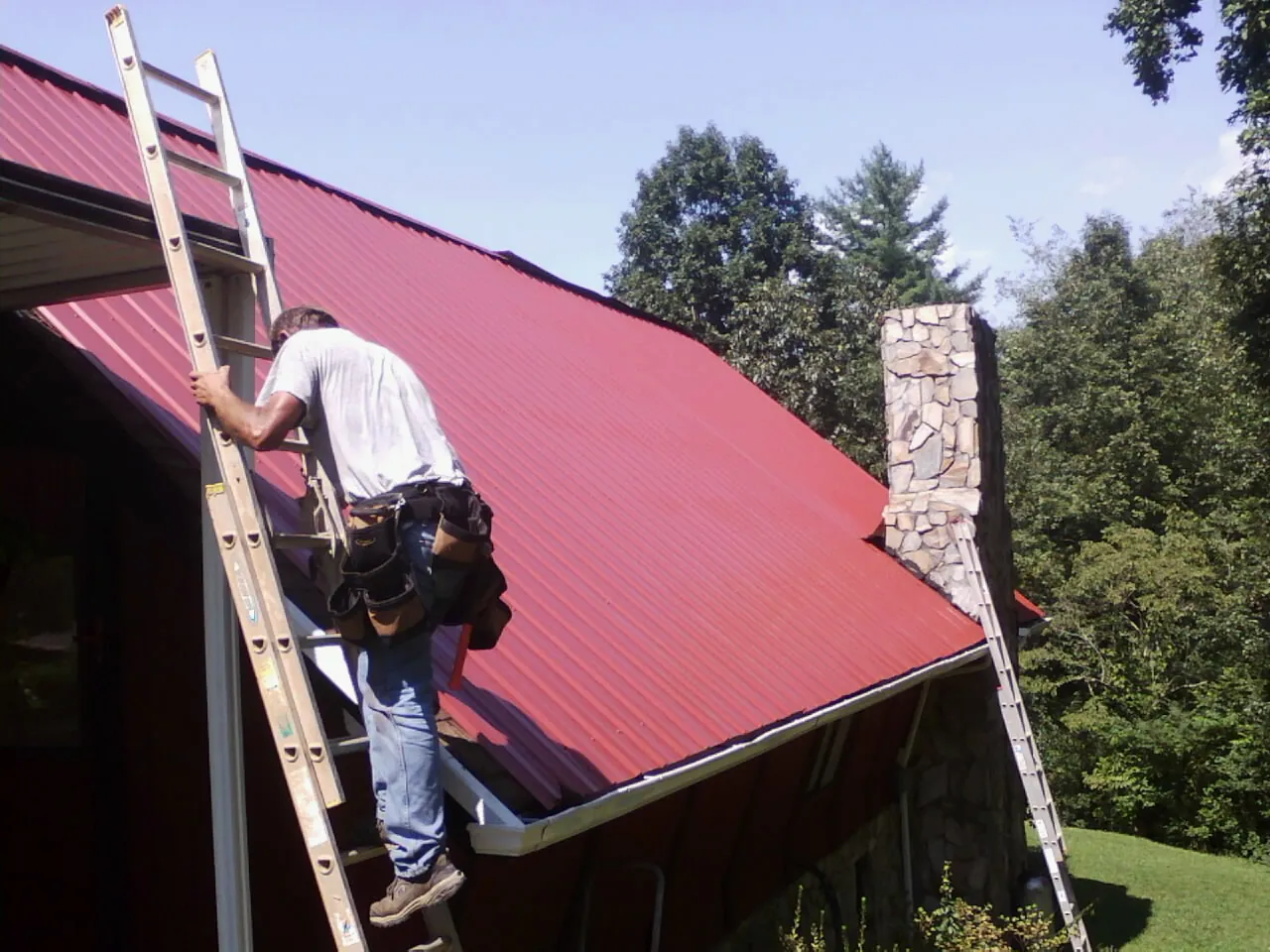 Roofer on a ladder beside a red metal roof and stone chimney — Soffit & Fascia Repair by Roofing Brothers in Linthicum Heights